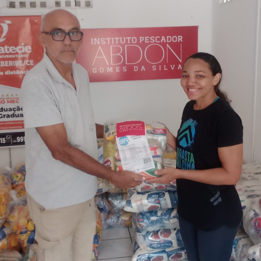 Homem entregando uma cesta básica de doação a uma mulher, ambos segurando o item, com várias cestas básicas empilhadas ao fundo e a placa do Instituto Pescador Abdon Gomes da Silva na parede.